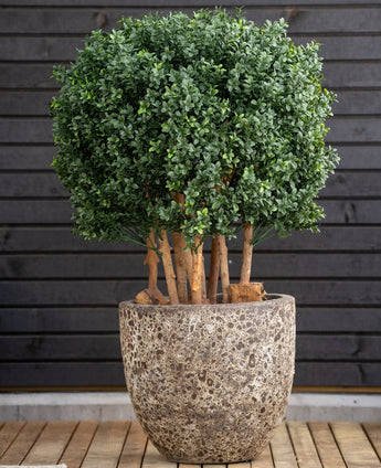 Artificial Boxwood Ball Wood Roman Style UV 90 cm, en rund bladgrøn plante med tykke stængler, står i en stor stenpotte på træ, mod en mørk vandret væg med træpaneler.