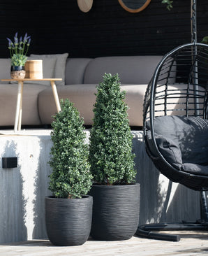 Twee Kunstplant Buxus Pyramide UV 120 cm staan op een houten dek voor een buitenzitje met een lichte bank, ronde tafel met lavendel en zwarte hangstoel.