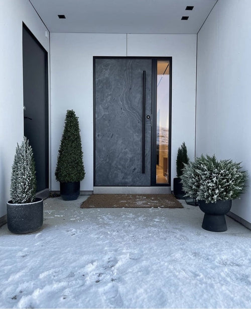 En moderne mørk hoveddør er indrammet af hvide vægge og flankeret af Artificial Plant Boxwood Pyramid UV 90 cm, som giver UV-beskyttelse. Let sne dækker jorden, mens disse stilfulde haveplanter skaber en ryddelig, minimalistisk entré.