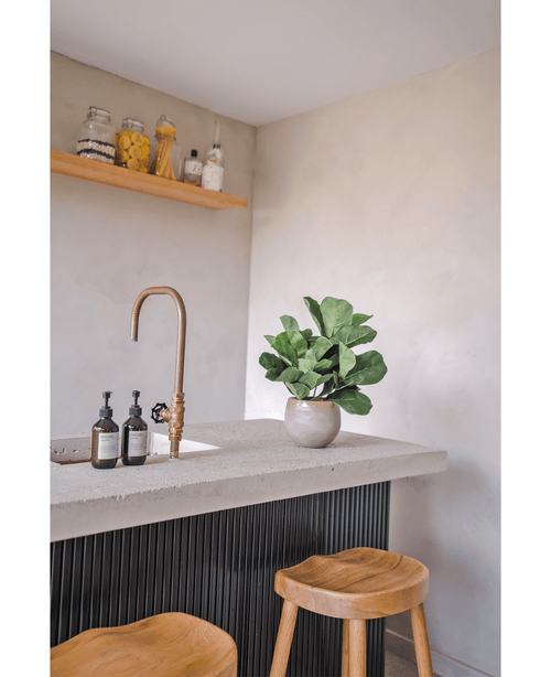 En moderne bordplade med en messinghane, to sæbedispensere, den kunstige tobaksplante Fiddle Leaf UV 45 cm potteplante og to træskamler. En flydende hylde med krukker og flasker er monteret på en lys væg.
