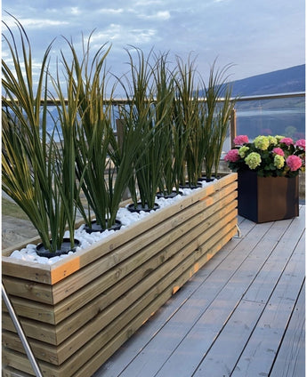 En plantekasse i træ med kunstgræsgrøn UV 120cm og hvide sten står på en balkon med glasrækværk; i nærheden, en sort firkantet potte med blomstrende lyserøde, grønne og hvide hortensiaer har udsigt over den maleriske udsigt.