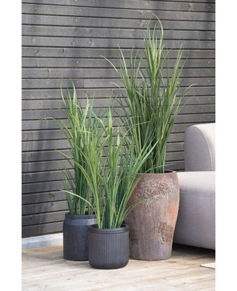 En kunstgræsgrøn UV 90 cm kunstig plante i en sort potte står på et trægulv ved siden af ​​en grå udendørs sofa, med en mørk træspaltevæg i baggrunden.