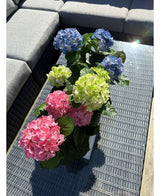 Artificial Hydrangea Green UV 35 cm planten står på et fletbord med glasplade ved siden af ​​en grå udendørs sofa, med realistiske grønne hortensiaer, der lyser op i sollys.