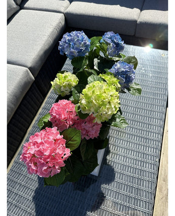 Artificial Hydrangea Green UV 35 cm planten står på et fletbord med glasplade ved siden af ​​en grå udendørs sofa, med realistiske grønne hortensiaer, der lyser op i sollys.