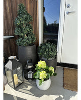 En Kunstig Hortensia Grøn UV 35 cm plante i en hvid potte med gulgrønne blomster står på verandaen nær en brun dør, ved siden af ​​to planter i en sort potte og to dekorative lanterner med stearinlys.