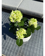 Artificial Hydrangea Green UV 35 cm, med lysegrønne blomster og mørkegrønne blade, står på et vævet gråt udendørsbord med en polstret bænk i baggrunden.