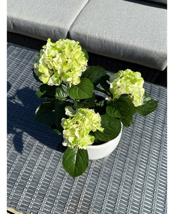 Artificial Hydrangea Green UV 35 cm, med lysegrønne blomster og mørkegrønne blade, står på et vævet gråt udendørsbord med en polstret bænk i baggrunden.