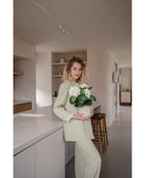 En kvinde med krøllet blond hår, klædt i et lysegrønt outfit, står i et moderne køkken med en Artificial Hydrangea White UV 35 cm plante i hænderne – med cremede hvide blomster – og smiler til kameraet.
