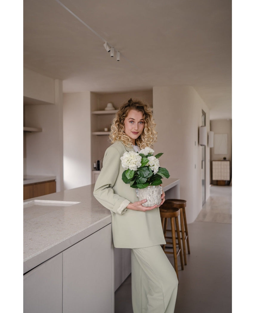 En kvinde med krøllet blond hår, klædt i et lysegrønt outfit, står i et moderne køkken med en Artificial Hydrangea White UV 35 cm plante i hænderne – med cremede hvide blomster – og smiler til kameraet.