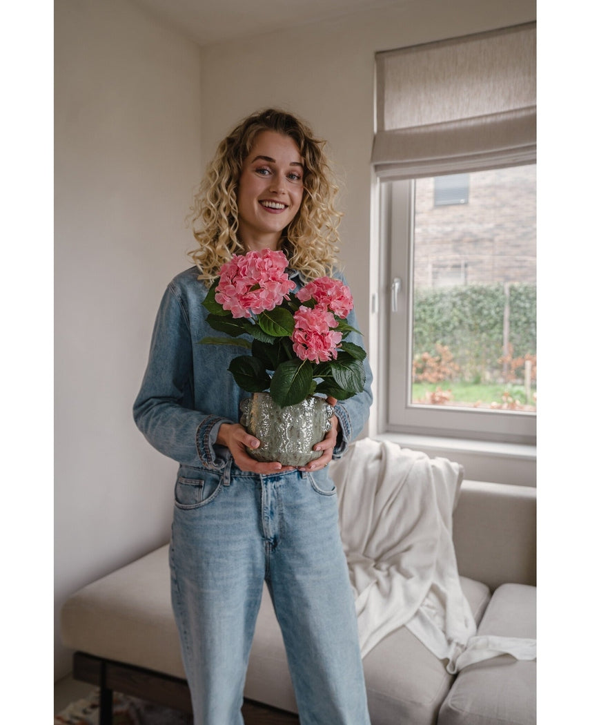 En kvinde med krøllet blond hår i denimjakke og jeans står indendørs og smiler, mens hun holder den Artificial Hydrangea Pink UV 35 cm plante. I baggrunden er der en sofa og et vindue.