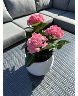 Den kunstige plante Hortensia Pink UV 35 cm, en hvid potte med levende lyserøde hortensiablomster, står på et vævet gråt udendørsbord med en grå sofa med puder i baggrunden, oplyst af sollys og bløde skygger.