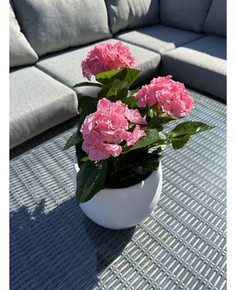 Den kunstige plante Hortensia Pink UV 35 cm, en hvid potte med levende lyserøde hortensiablomster, står på et vævet gråt udendørsbord med en grå sofa med puder i baggrunden, oplyst af sollys og bløde skygger.