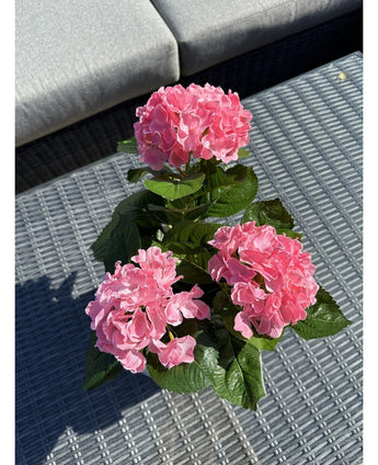 Den Kunstige Plante Hortensia Pink UV 35 cm, med tre lyserøde hortensiablomster og grønne blade, står på et vævet gråt udendørsbord, med en grå sofa med puder i baggrunden.