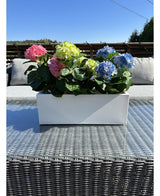 Den Kunstige Plante Hortensia Pink UV 35 cm, en rektangulær hvid plantekasse med lyserøde hortensiaer, er placeret udenfor på et fletbord med en glasplade, grå hynder og en blå himmel i baggrunden.