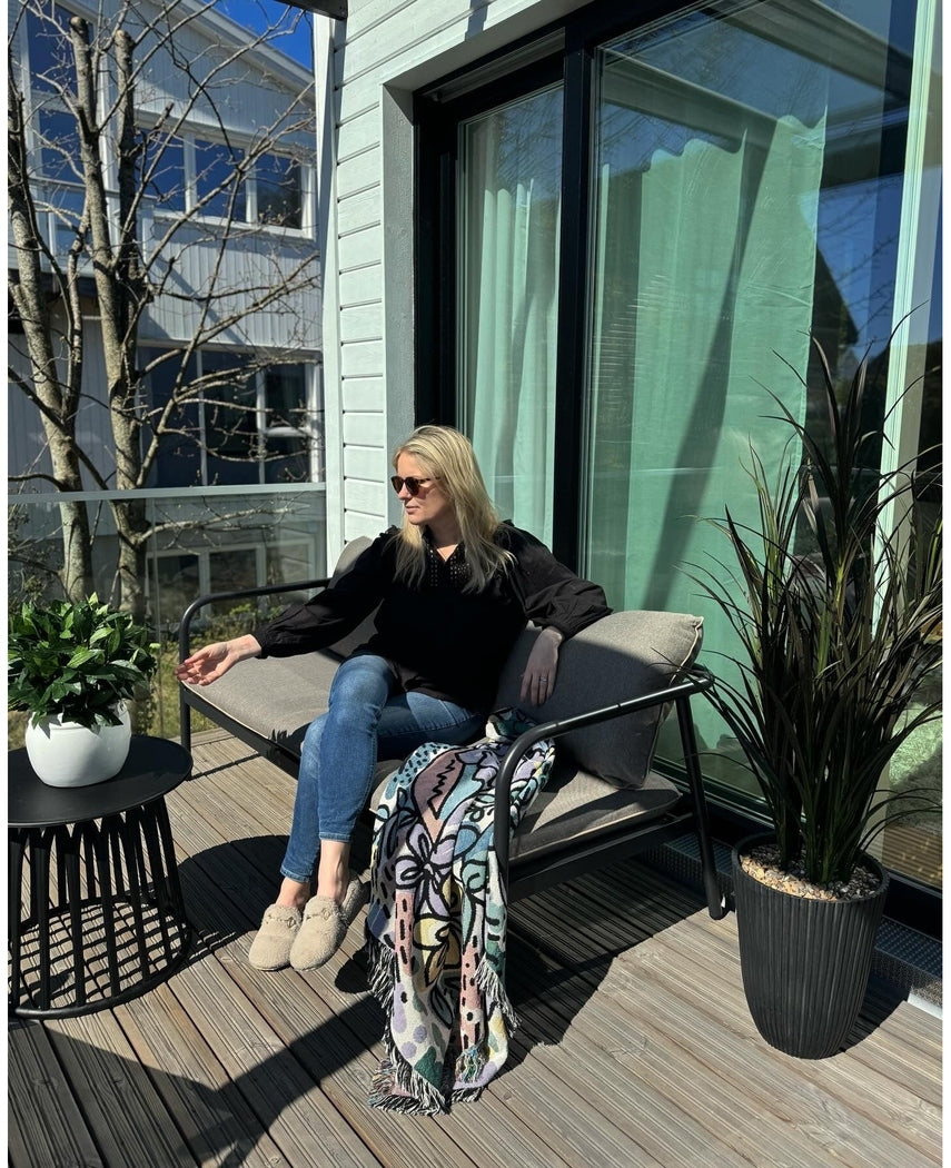 Een vrouw met zonnebril, zwart topje en blauwe jeans ontspant op een kussensstoel op een zonnig balkon met een drankje in de hand. De ruimte is voorzien van de Kunstplant Laurier UV d33 cm, een levendig decor, een kleurrijke deken en een kleine tafel met planten.