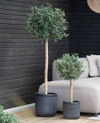 Twee Kunst olijfboom UV potten van 150 cm met ronde groene bladeren staan in zwarte potten op een houten vloer bij een beige buitenbank, tegen een donkere houten muur. Beide bomen zijn UV-bestendig.