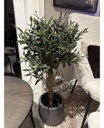 Det kunstige oliventræ UV 85 cm, en kompakt og letplejet kunstig plante med mørkegrønne blade og sorte oliven, står indendørs ved siden af ​​et rundt træbord og en lænestol med hvid tekstur.