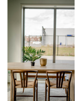 Et træbord med to stole, en kunstig palmesalon UV 60 cm i en krukke og en guldvase er placeret i nærheden af ​​et stort vindue med udsigt over et industriområde med bygninger og et hegn.
