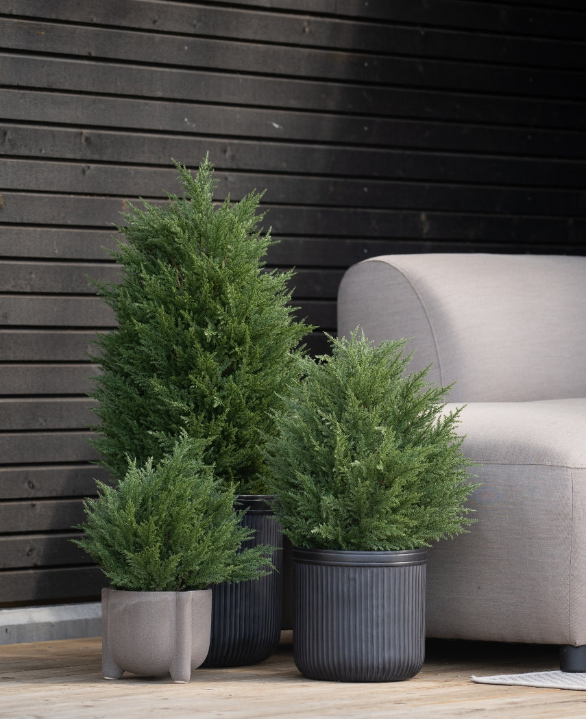 Tre kunstige nåletræer deluxe UV 60 cm kunstige buske står på et træterrasse nær en lysegrå udendørs bænk og en mørk trævæg - perfekt til at skabe en grøn have med kunstige planter udenfor.