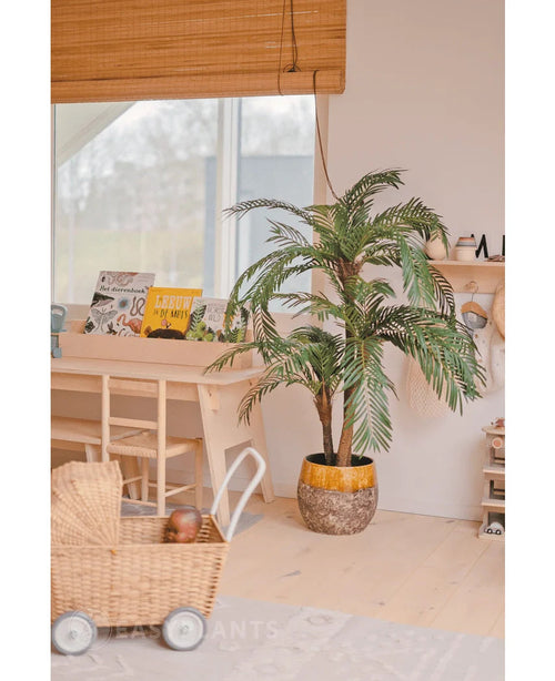 I et hyggeligt, lyst rum er Artificial Palm Cycas Classic 135 cm placeret i en stilfuld plantekasse, en flettet legetøjsvogn, træmøbler og børnebøger nær et vindue med et bambus-rullegardin.