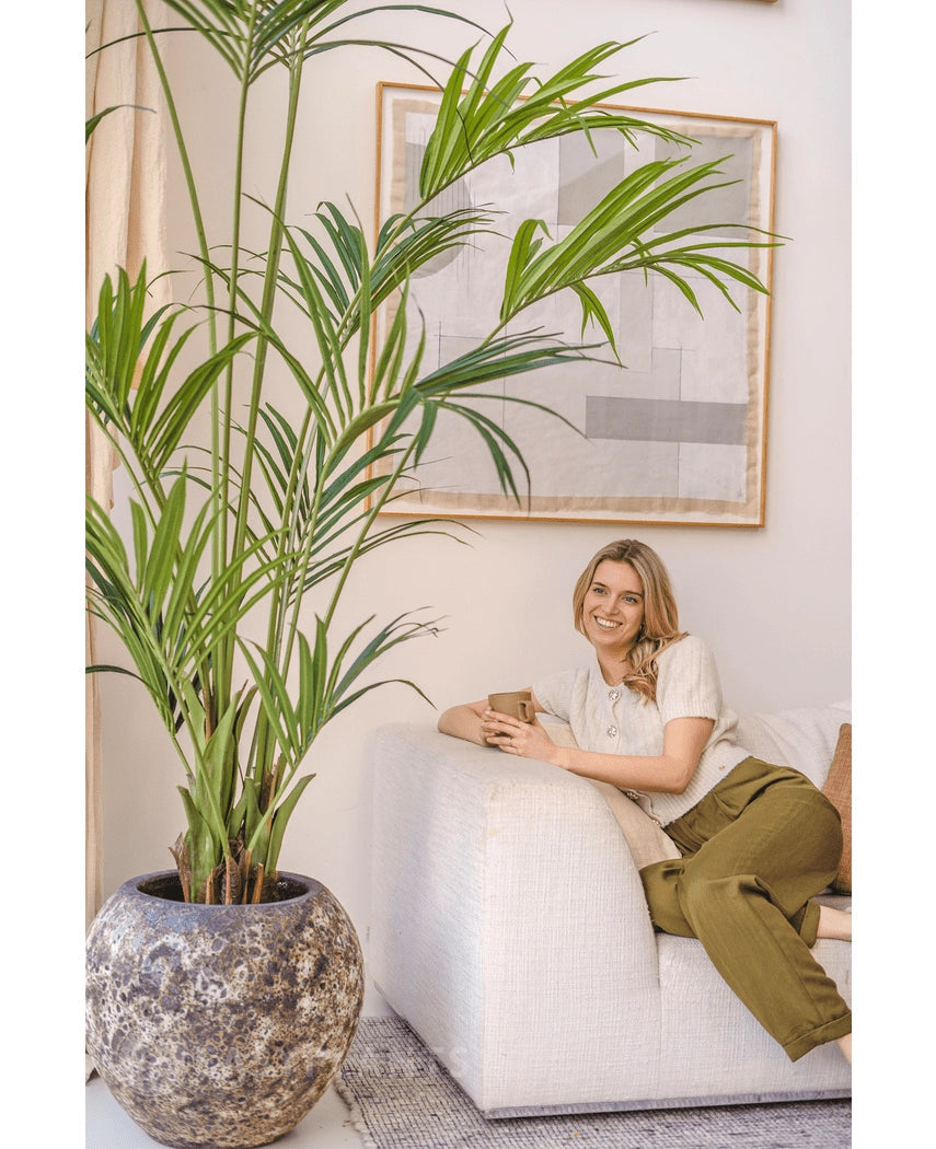 En kvinde med lyst hår, i en cremet top og olivengrønne bukser, sidder i en hvid sofa og smiler og holder et krus. Den kunstige Kentia Palm 220 cm forstærker den tropiske atmosfære i det lyse, hyggelige rum med abstrakt vægkunst.