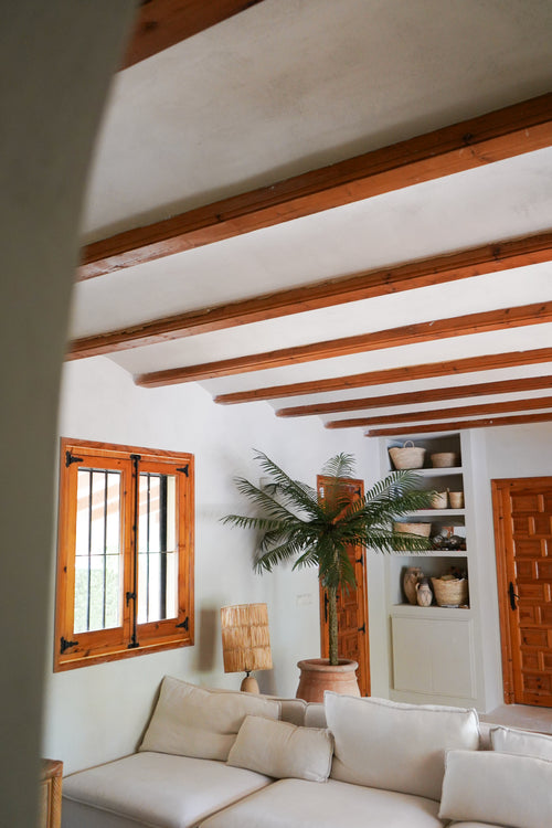 En hyggelig stue med hvide vægge, træbjælker, en hvid sektionssofa, Artificial Palm Phoenix De Luxe 210 cm, trævindue og dør, indbyggede hylder med kurve og keramik og naturligt lys, der kommer ind gennem vinduet.
