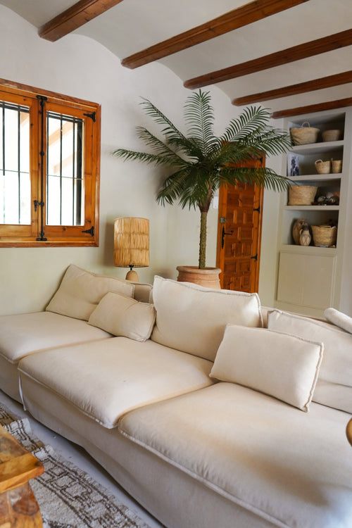 En hyggelig stue med en stor cremefarvet sofa, Artificial Palm Phoenix De Luxe 210 cm i hjørnet, en trælampe, indbyggede hylder med kurve, loftsbjælker i træ og et vindue med trærammer.