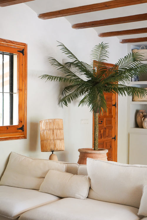 En hyggelig stue med en hvid sofa, en rattan bordlampe, Artificial Palm Phoenix De Luxe 210 cm i en potte, træbjælker i loftet, et vindue med trærammer og en trædør i baggrunden.