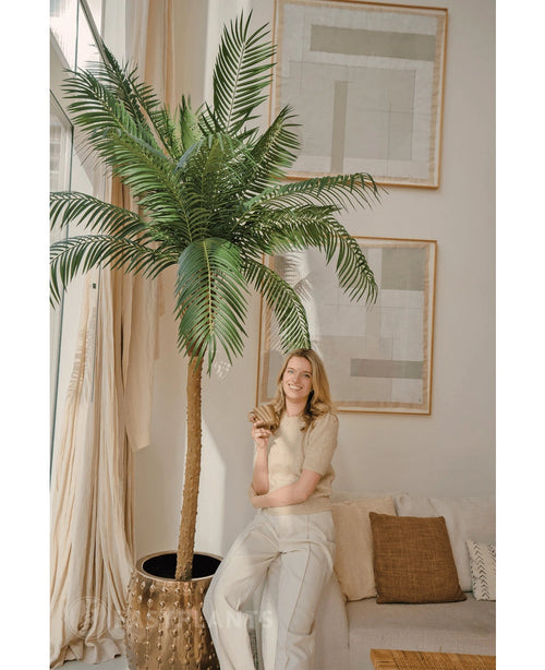 En smilende kvinde står ved siden af ​​Artificial Palm Phoenix De Luxe 250 cm i en lys, stilfuld stue med neutral indretning og stor abstrakt kunst. Hun holder en plante og læner sig afslappet op ad en beige sofa med puder.