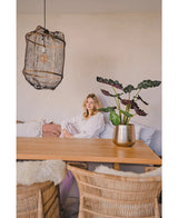 En kvinde i en hvid bluse sidder ved et spisebord af træ med kurvestole. Det hyggelige, blødt oplyste rum har kunstplanten Alocasia UV 67 cm og en struktureret hængelampe som indretning.