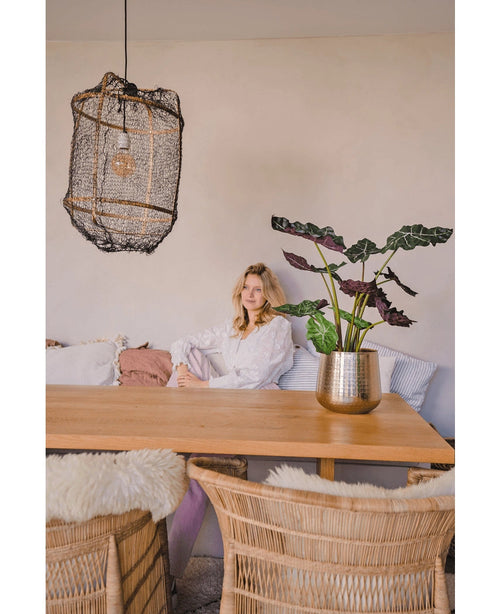 En kvinde i en hvid bluse sidder ved et spisebord af træ med kurvestole. Det hyggelige, blødt oplyste rum har kunstplanten Alocasia UV 67 cm og en struktureret hængelampe som indretning.