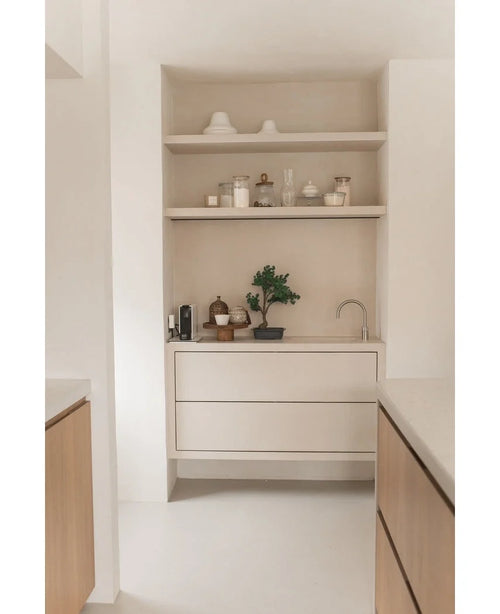 Minimalistisk køkkenhjørne med lyse træhylder med potter, vaser, indretning, en bordplade med små hvidevarer og den Kunstige Bonsai Juniper 40 cm potteplante, plus indbyggede skuffer og en lille hane - alt i neutrale toner.