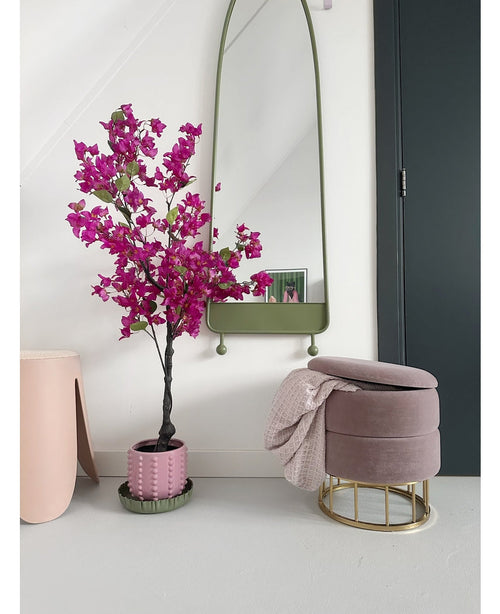 Et moderne interiør med Artificial Bougainvillea 125 cm Lilla i en dekorativ potte, et højt spejl med grøn ramme, en pink fodskammel med guldbund og pink tæppe og en mørk dør mod en sprød hvid væg.