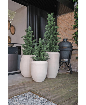 Drie hoge, witte plantenbakken met Kunstplant Conifeer UV 90cm staan op een houten terras naast een modern gebouw. Een zwart keramisch rooster staat vlakbij tegen een stenen muur met wijnranken.
