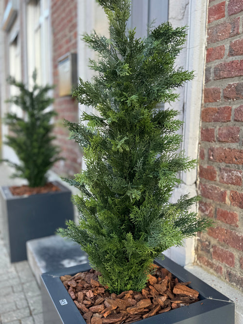 Et nærbillede af det kunstige nåletræ UV 120 cm, en grøn kunstig nåletræsplante i en rektangulær sort plantekasse med brun barkbark, vist på et fortov nær en murstensbygning; en anden sådan plantekasse er i baggrunden.