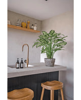 Een moderne keuken met een betonnen aanrechtblad, twee houten krukjes en een Kunstplant Dieffenbachia 75 cm bij de messing kraan, plus zeepdispensers en een houten plank met potten tegen een lichtgrijze muur.