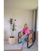 En kvinde i en lys pink top og blå jeans sidder barfodet på en moderne sort og flet stol ved siden af ​​den kunstige plante Dracaena Fragnans 150 cm i et lyst, minimalistisk rum med hvide vægge og gulv.