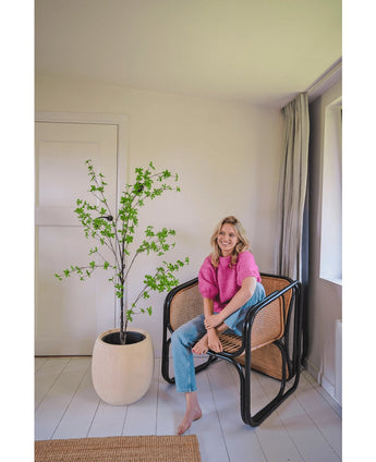 Een vrouw in een roze topje en blauwe spijkerbroek zit blootsvoets op een moderne rieten stoel naast de Kunstplant Enkianthus 150 cm in een lichte, minimalistische kamer met witte muren en lichte vloerbedekking.