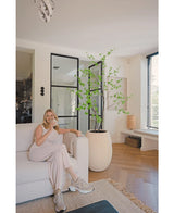 En kvinde i let loungewear sidder i en beige sofa og smiler med en kop kaffe i en moderne stue med trægulve, gulv til loft vinduer og den Kunstige Enkianthus 180 cm som en stor potteplante.