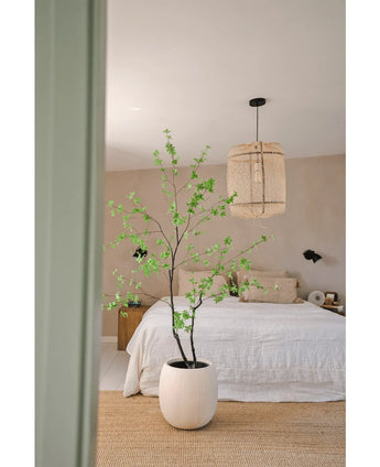 Et minimalistisk soveværelse med Artificial Enkianthus 180 cm planten som stor potteplante, et bed med hvidt sengetøj, en vævet hængelampe, beige vægge og et naturfibertæppe for en enkel og luftig atmosfære.
