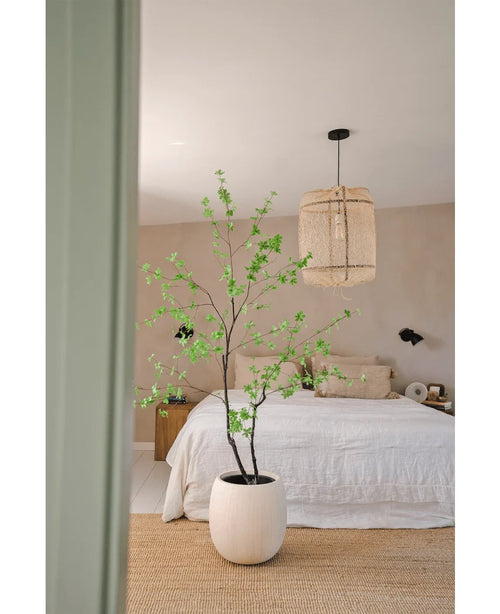 Et minimalistisk soveværelse med Artificial Enkianthus 180 cm planten som stor potteplante, et bed med hvidt sengetøj, en vævet hængelampe, beige vægge og et naturfibertæppe for en enkel og luftig atmosfære.