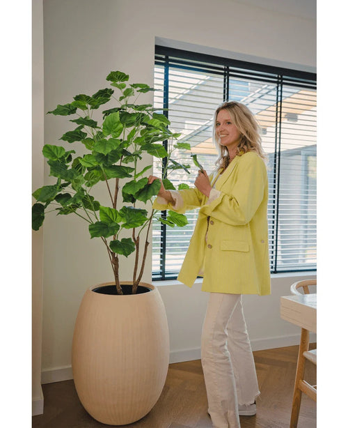 En kvinde i gul blazer og hvide bukser smiler, mens hun passer den kunstige planteficus 150 cm i et moderne, veloplyst rum med trægulv og store vinduer med persienner.