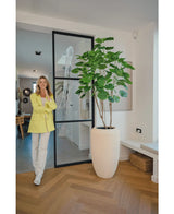Een vrouw in een gele blazer en witte broek glimlacht naast de Kunstplant Ficus 180 cm in een moderne, lichte kamer met houten vloer en minimalistisch decor.