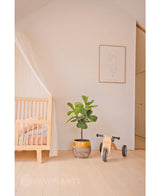 Een minimale kinderkamer met een licht houten ledikant, een doorschijnende luifel, Kunstplant Ficus Tabaksplant Deluxe 90 cm, houten speelgoed met het label Mia en een minimalistische lijnkunst ingelijste print op een effen witte muur.
