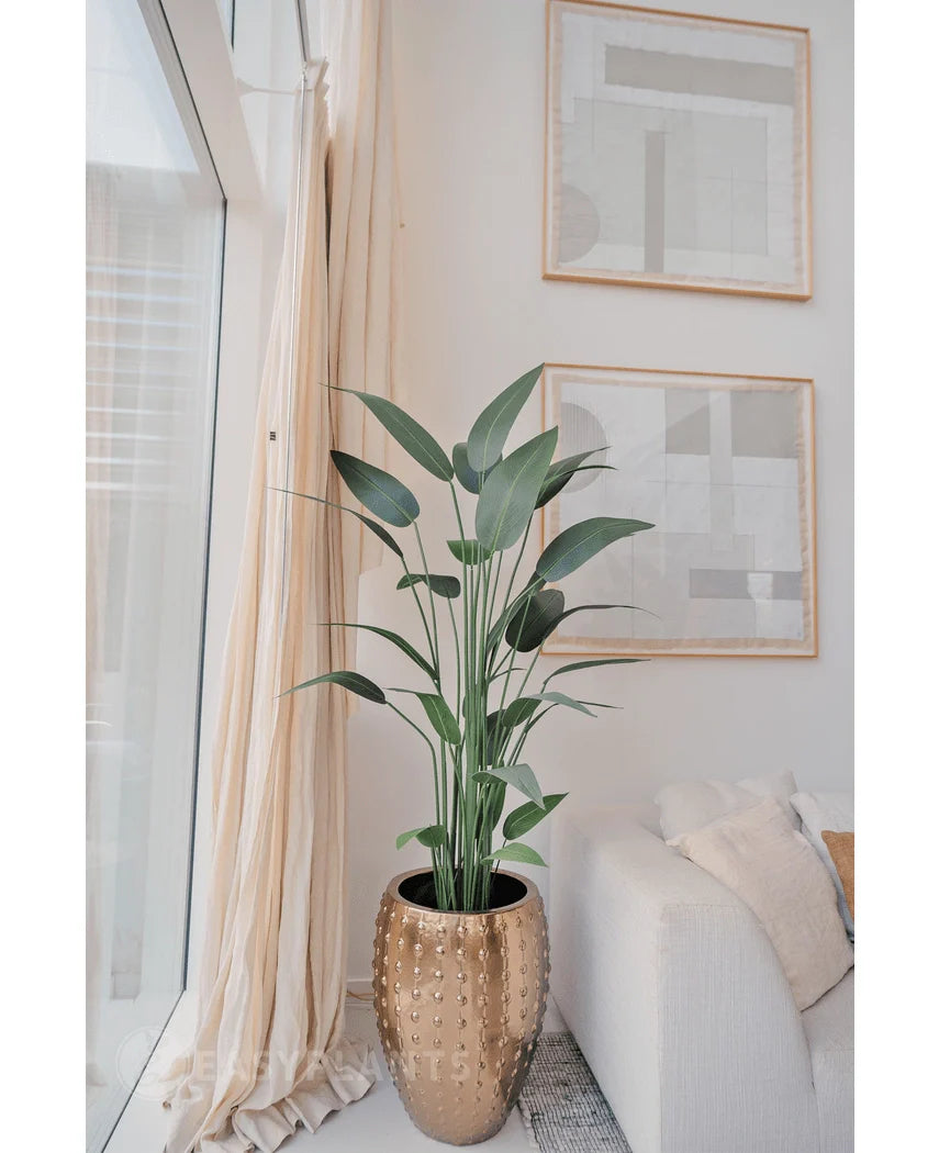 De Kunstplant Heliconia 180 cm in een goudkleurige plantenbak staat bij een raam met transparante gordijnen, naast een witte bank. Twee abstracte kunstwerken hangen erboven - perfect voor wie een kunstplant wil kopen.