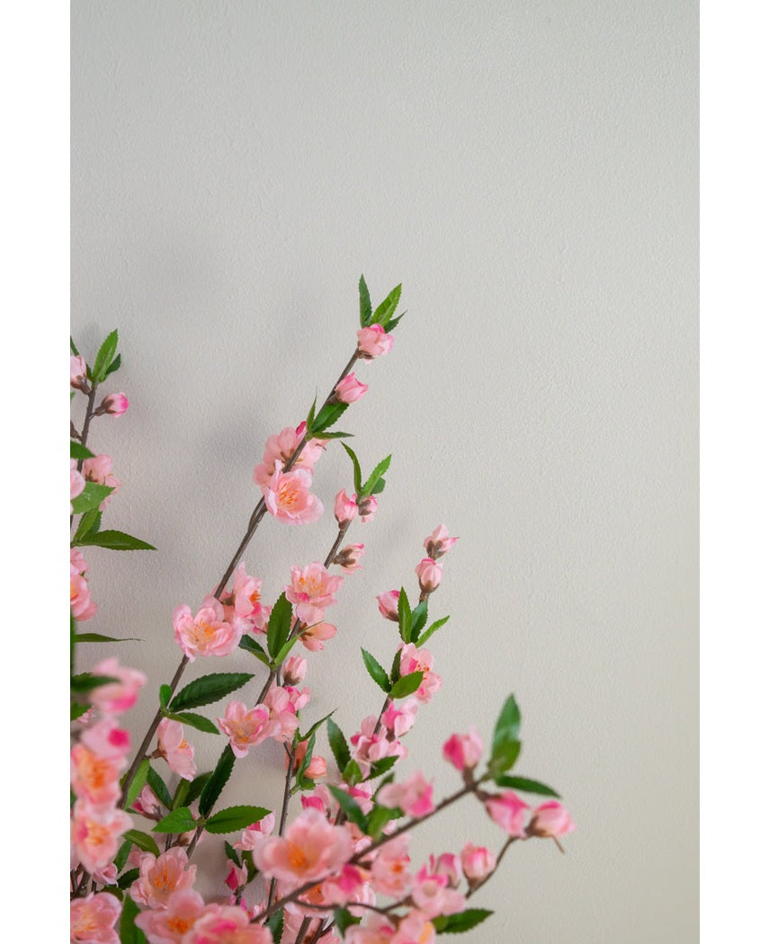 De Kunstplant Kersenbloesem 120 cm roze roze kersenbloesemtakken met groene bladeren die linksonder uitsteken op een effen, lichtbeige achtergrond.