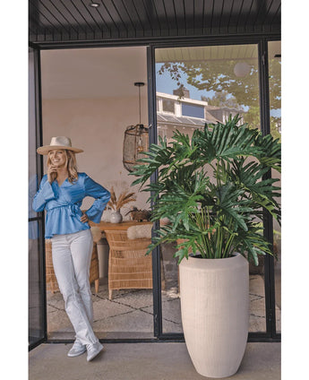Een vrouw in een blauwe blouse, witte broek en hoed lacht bij een glazen deur naast de Kunstplant Philodendron 125 cm in een beige pot. Rieten stoelen en decor zijn zichtbaar in de kamer.