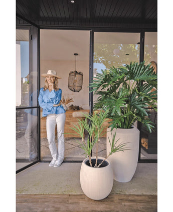 Een vrouw met hoed, blauwe blouse en witte broek leunt tegen een glazen deur. Op de voorgrond staat de Kunstplant Philodendron 125 cm. Binnenin is een gezellige, moderne leefruimte te zien.