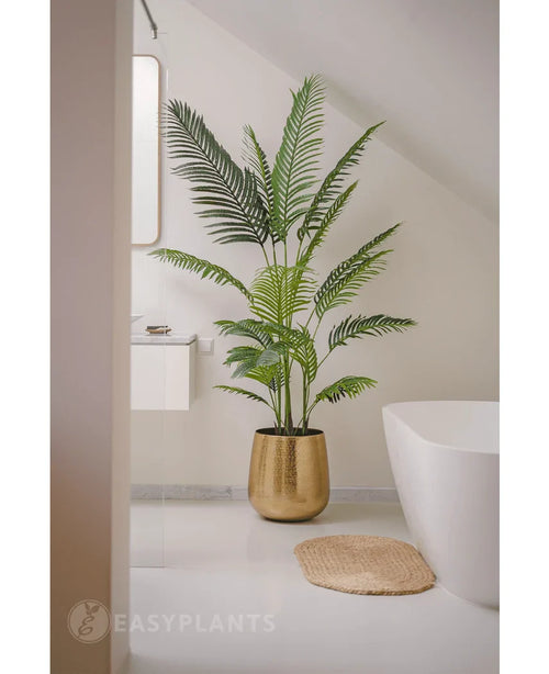 Kunstplanten Phoenix 180 cm i guldkrukke står ved siden af ​​et hvidt badekar og håndvask i et moderne minimalistisk badeværelse med hvide vægge og gulv. I nærheden ligger en rundvævet måtte på gulvet.