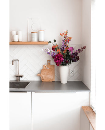 Et moderne hvidt køkken med en grå bordplade, en vask, et skærebræt i træ og en hvid vase fyldt med Kunstig Blomsterhortensia 55 cm mørk pink. Hvide kopper og gryder står på en træhylde over disken.